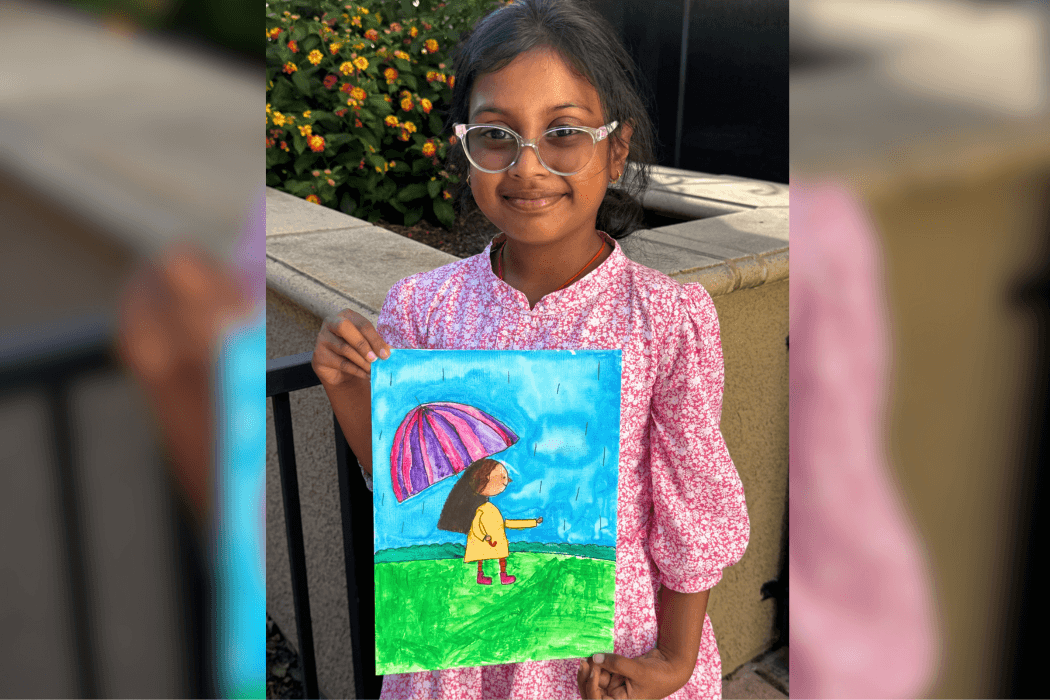 Shloka holding her Colorful Umbrella acrylic painting of a girl in a yellow raincoat under a pink-and-purple umbrella.